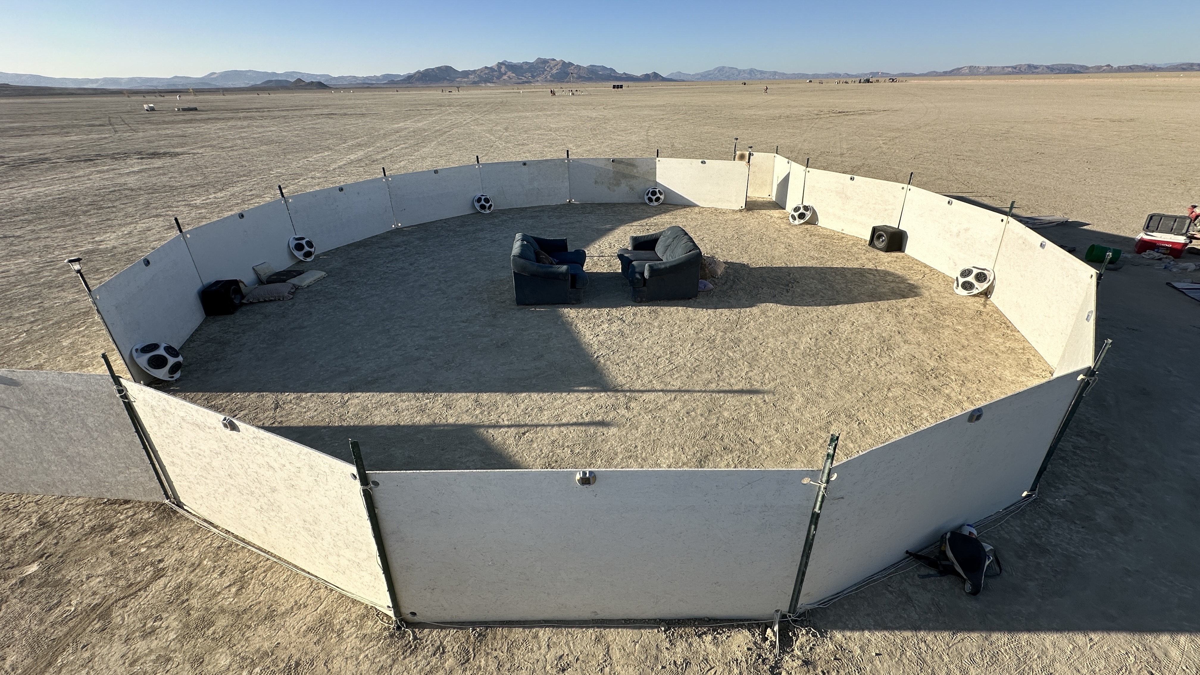 an image captured from above of a circle of low white walls surrounding two couches and a sound installation in the desert in daylight, near sunset