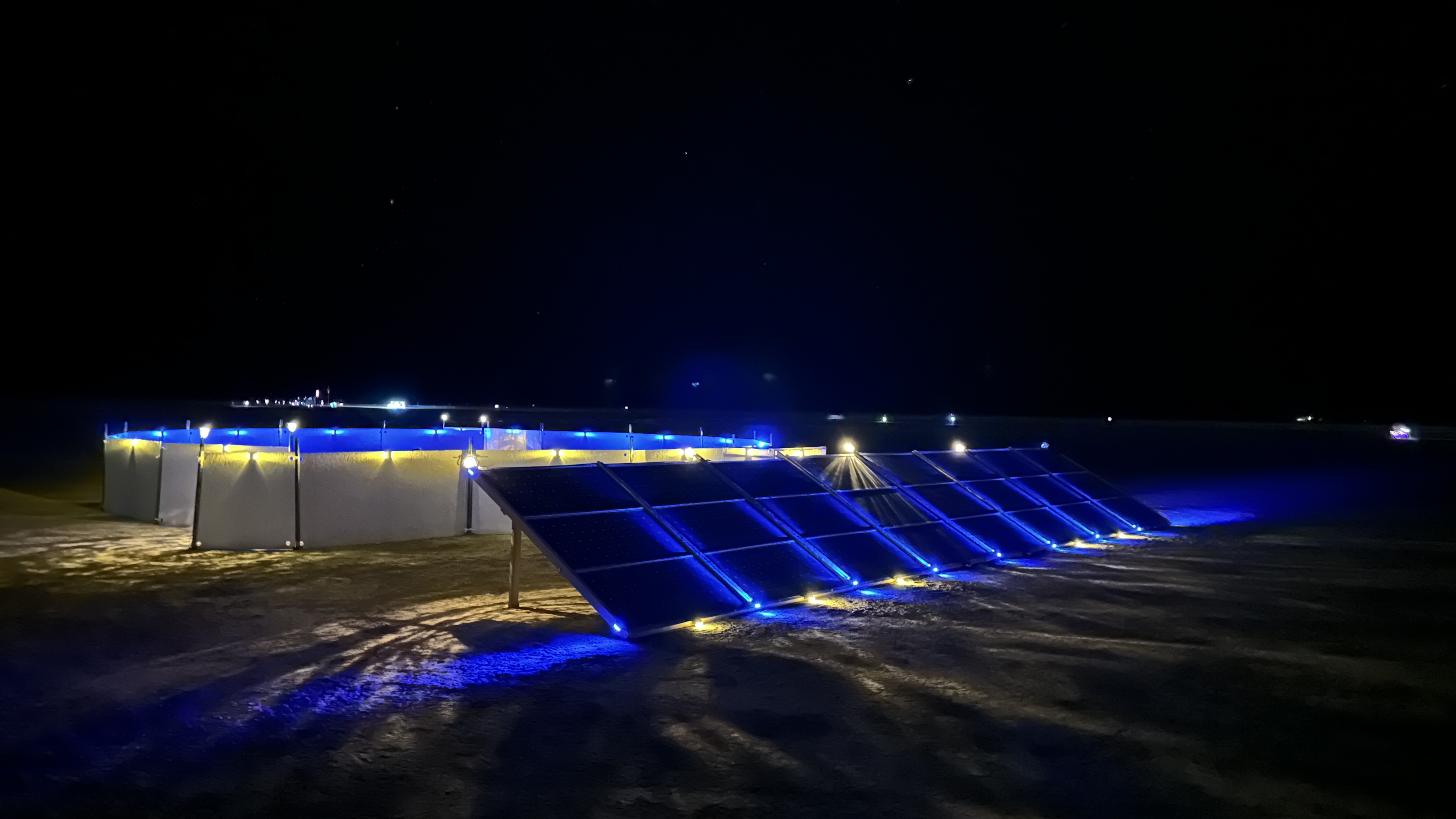 a circular sound installation in the desert with a large bank of prominent solar panels, lit up by white and blue LED lights at night