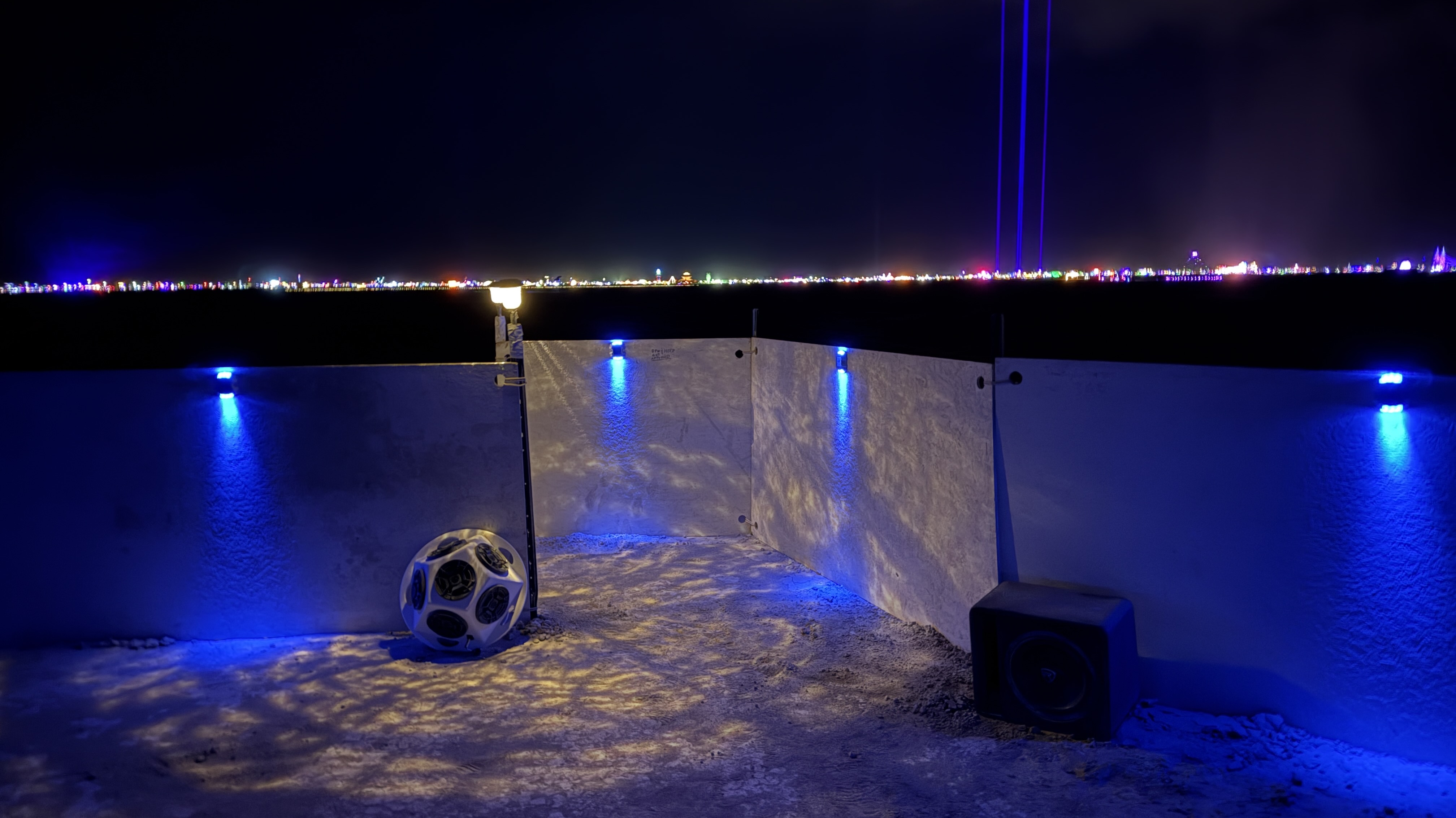 the interior side of an entrance to a circular sound installation in the desert at night, with the play of blue and white solar lights reflecting off of its surfaces and a hemisphere-shaped speaker leaning against the wall