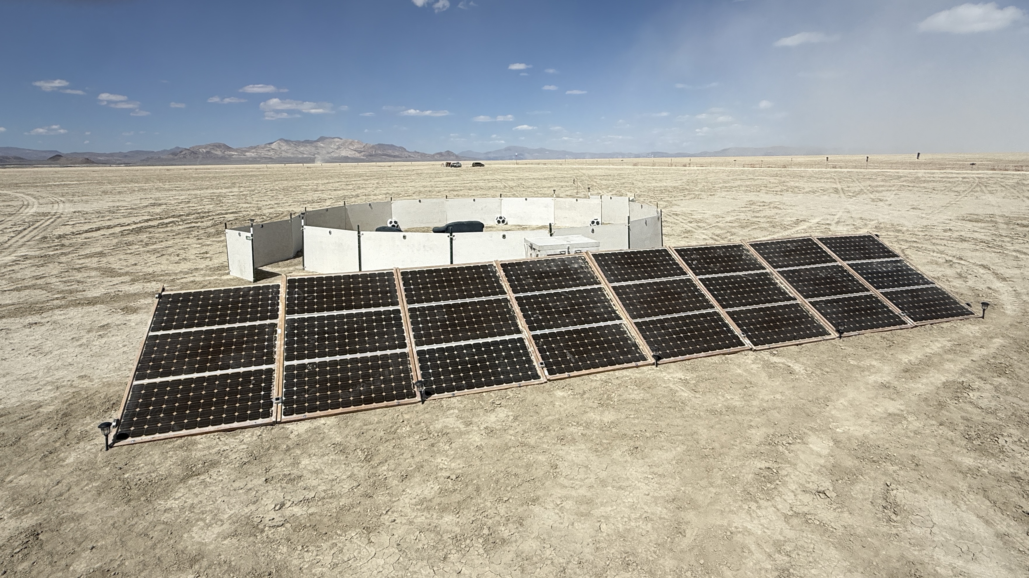A ring of low white walls sits behind a row of solar panels, in a desert setting
