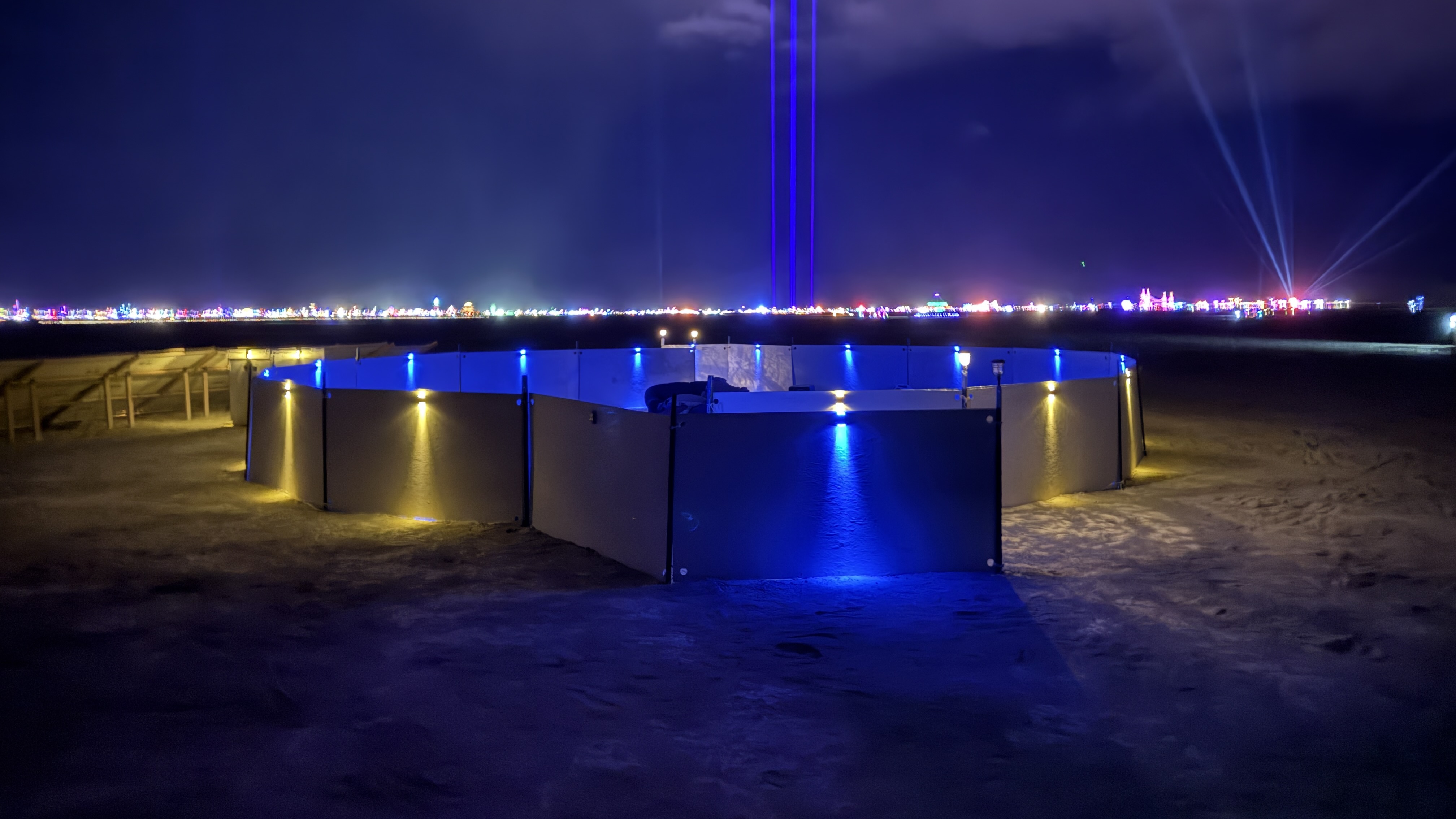 The Unpopular Music installation, a ring of low white walls adorned with solar-powered lights, sits on the ground before us with the nighttime lights of Black Rock City in the background